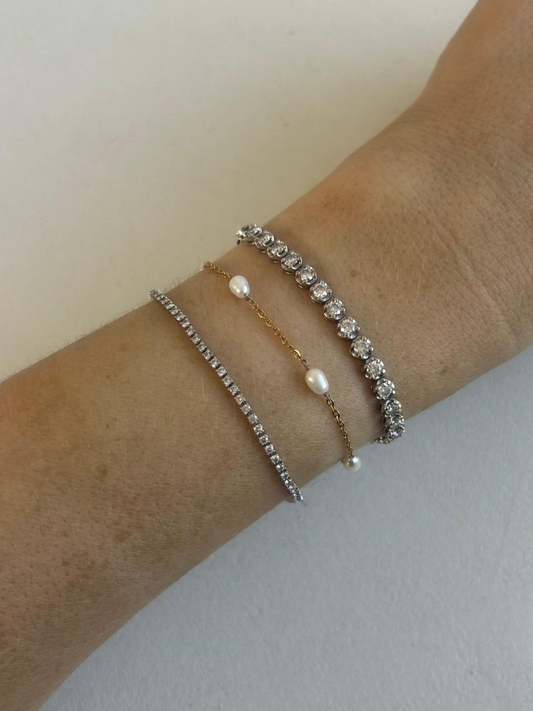 Three delicate bracelets stacked on a person's wrist: a silver tennis bracelet with clear stones, a gold chain bracelet with a pearl bead, and another silver stone bracelet. By Nicola and Coco & Lola Exclusive