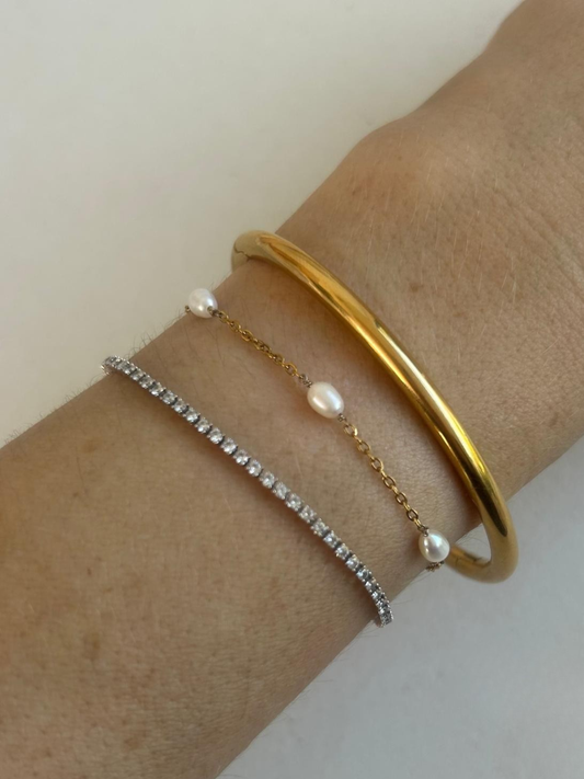 Three delicate bracelets worn on a wrist: a polished gold bangle, a silver tennis bracelet with clear stones, and a thin gold chain bracelet with white pearls. By Nicola and Coco & Lola Exclusive
