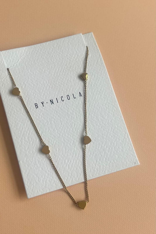 A gold necklace with small geometric charms displayed on a white textured card printed with the brand name 'BY-NICOLA', set against a solid peach background. Exclusive to Coco & Lola and By Nicola