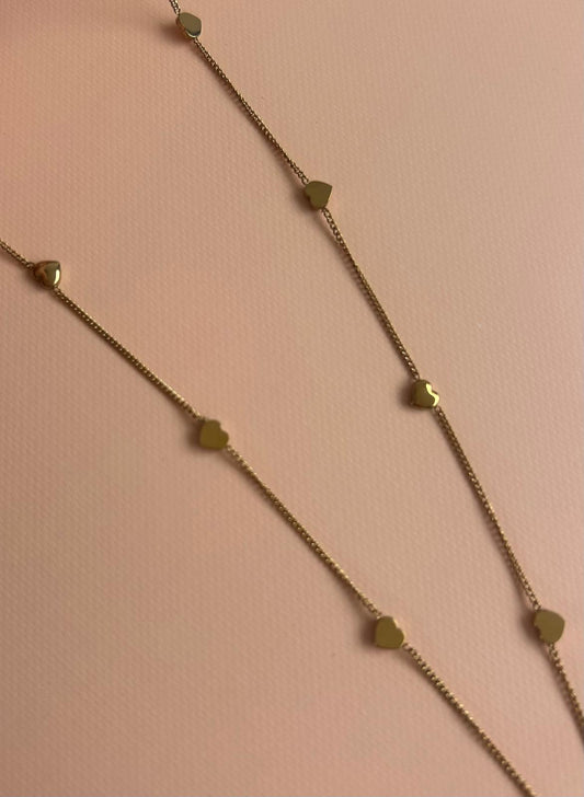A delicate gold chain necklace featuring small, spaced-out geometric heart-shaped charms, laid out diagonally on a soft pink textured background. Exclusive to Coco & Lola and By Nicola 